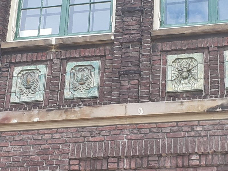 Ariensplein 3 siertegels Zaadbollen (katoen) en Spinnen (het bewerken) boven ingang vroegere Hogere Textielschool.jpg