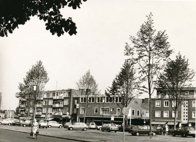 Raadhuisstraat 16 hoek  H J van Heekplein met bomen en parkeerterrein met geparkeerde auto's op achtergrond woonwinkelpanden, o.a. Anny Keyser, Arke en de winkel van Hollenkamp 1973.jpeg